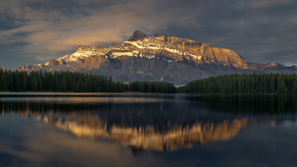 Mountain reflected in a lake with a forested area at the base under a cloudy sky.
