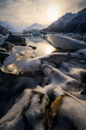 Frozen lake with ice and snow on rocks, surrounded by mountains under a cloudy sky.
