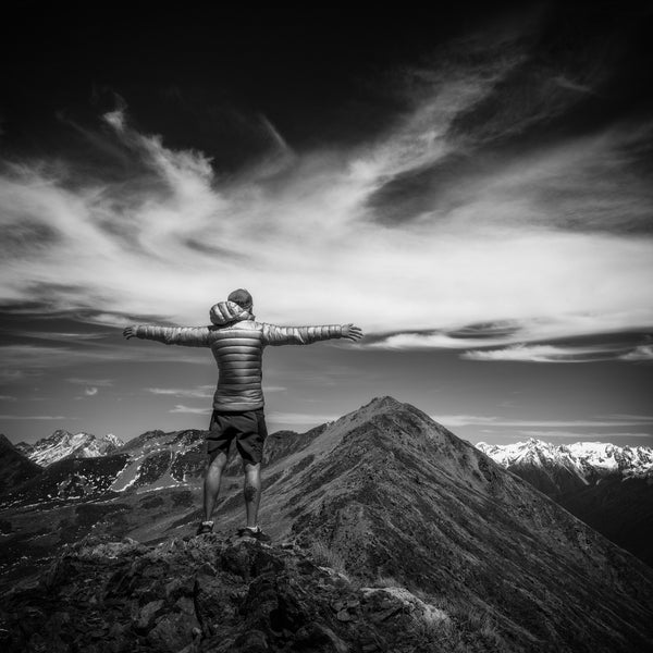 Jamil Slim standing on a mountain top with arms outstretched, surrounded by a dramatic sky and mountain range.